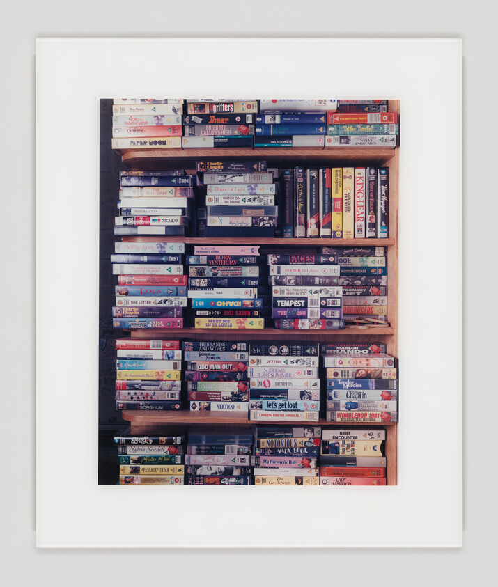 A framed photograph displaying a collection of vintage VHS tapes neatly arranged on shelves, with various movie titles visible on the spines.