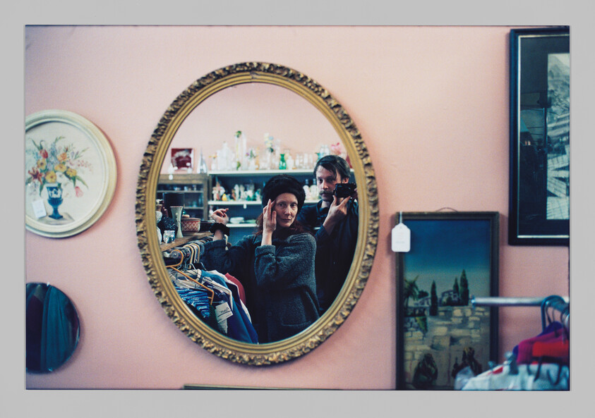 A woman adjusts a hat in a round mirror while a man photographs them in a thrift store.