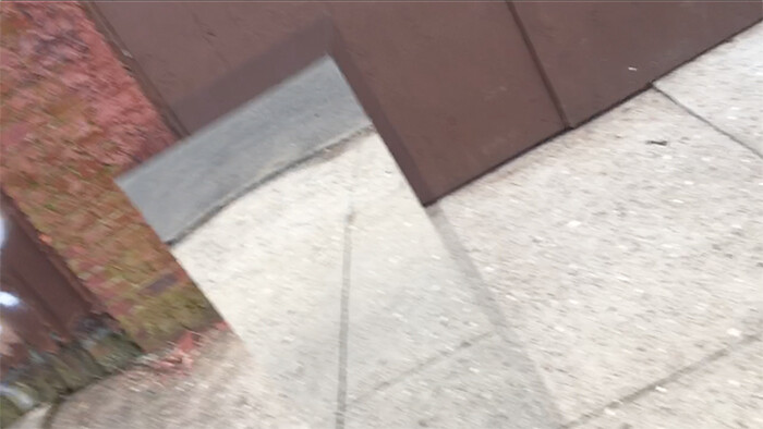 A rectangular mirror leans on a sidewalk, reflecting the pavement and a brown fence.