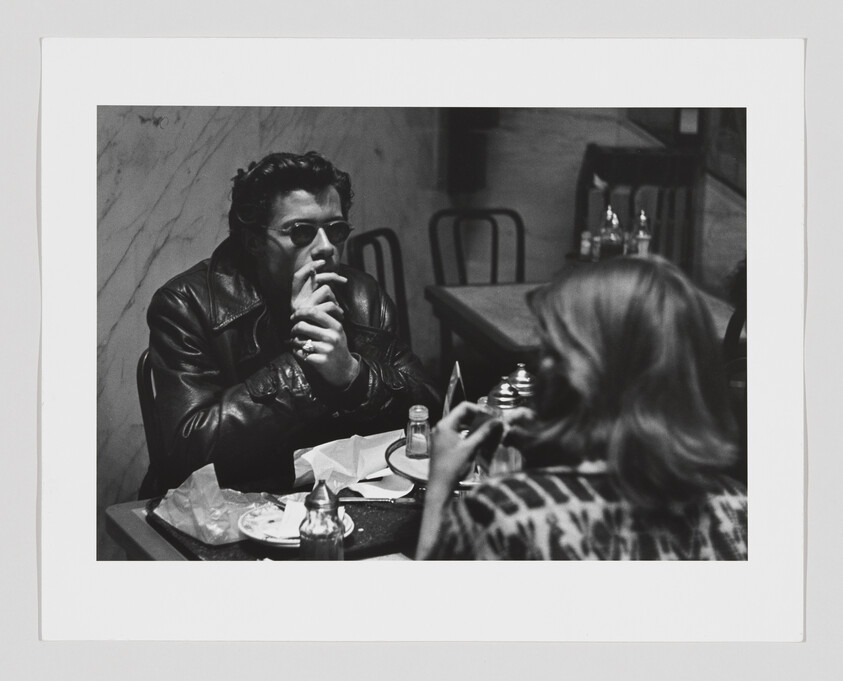 Man in a leather jacket and sunglasses smokes while talking with a woman at a diner table.