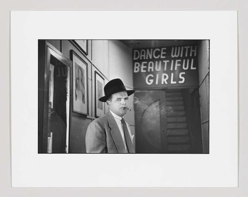Man in a suit and fedora smokes a cigar near a sign reading "Dance with beautiful girls."
