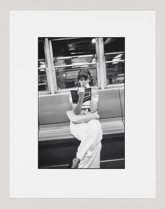 A black and white photograph of a woman sitting on a subway bench, engrossed in reading a book with the title "Ulysses" by James Joyce. She is wearing a striped top and white pants, with her legs crossed, and the subway car is in motion, as seen through the blurred windows behind her.