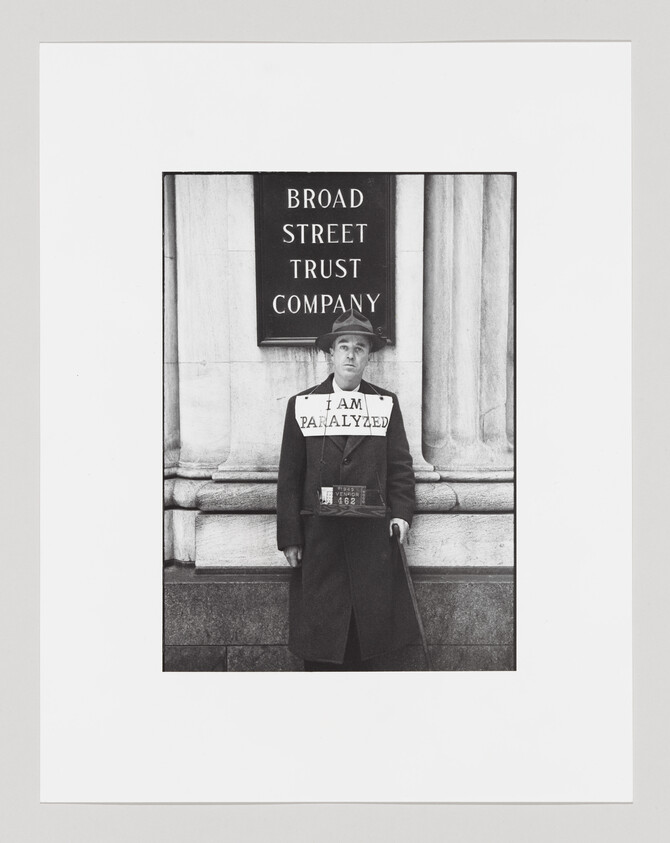 A man wearing a sign reading "I am paralyzed" stands with a cane in front of a bank.