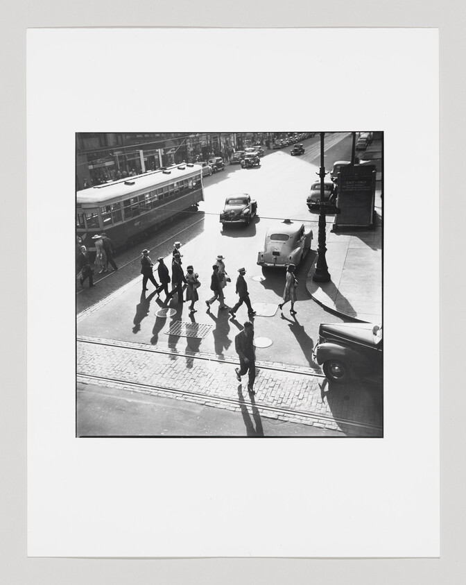 High-contrast black and white photograph capturing a group of people crossing a street in a city, with vintage cars and a tram in the background, viewed from an elevated angle.