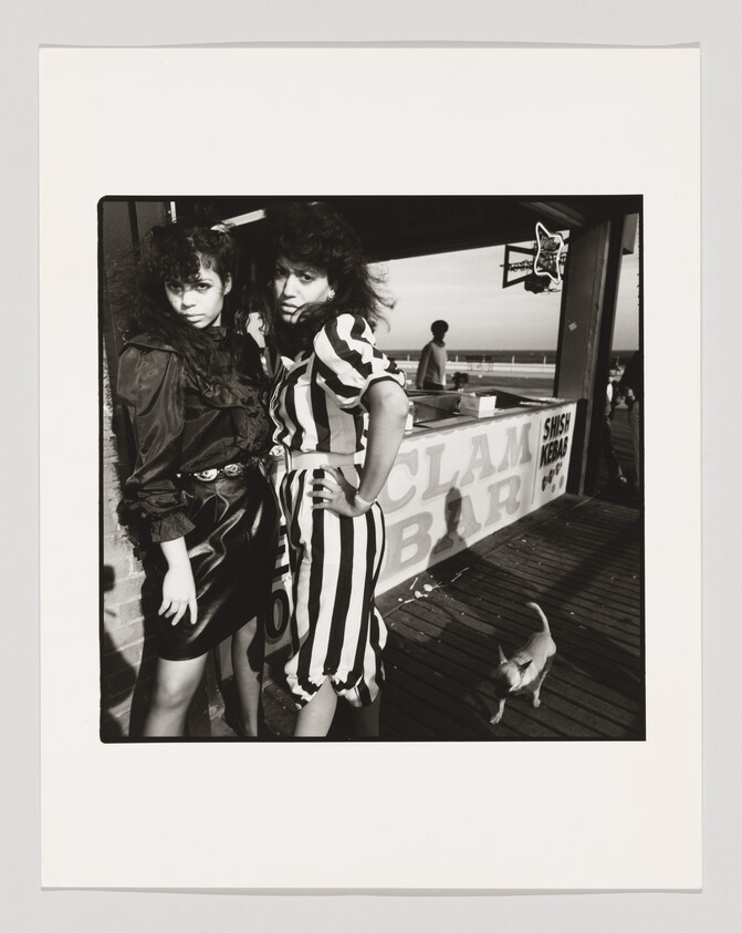 Two young women stand confidently by a boardwalk clam bar stall while a small dog walks nearby.