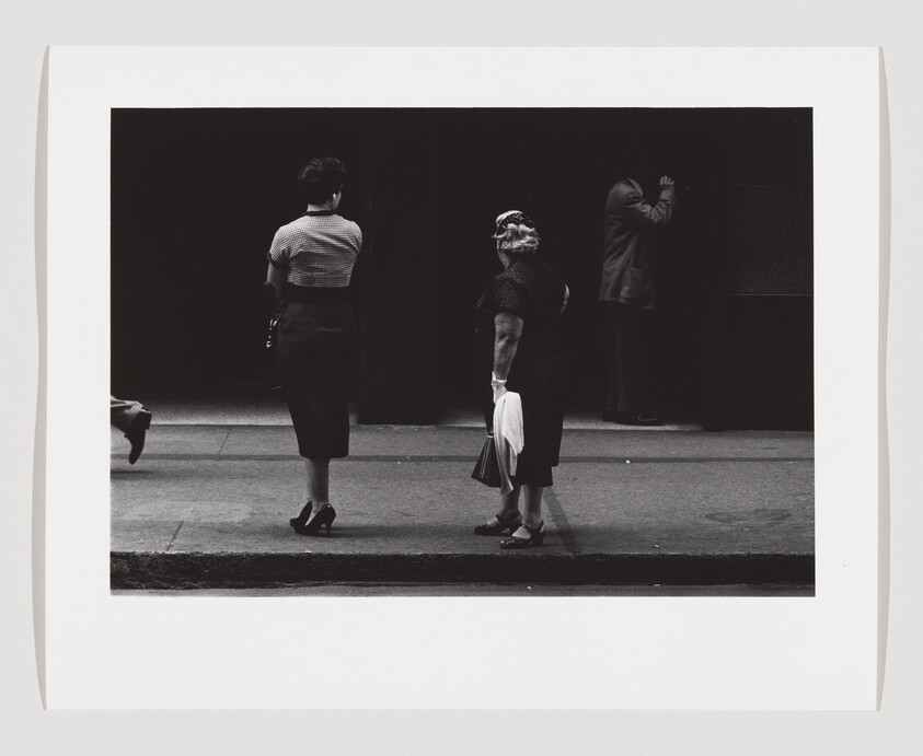 Two women wait on a city sidewalk while a man leans against a wall in the background.