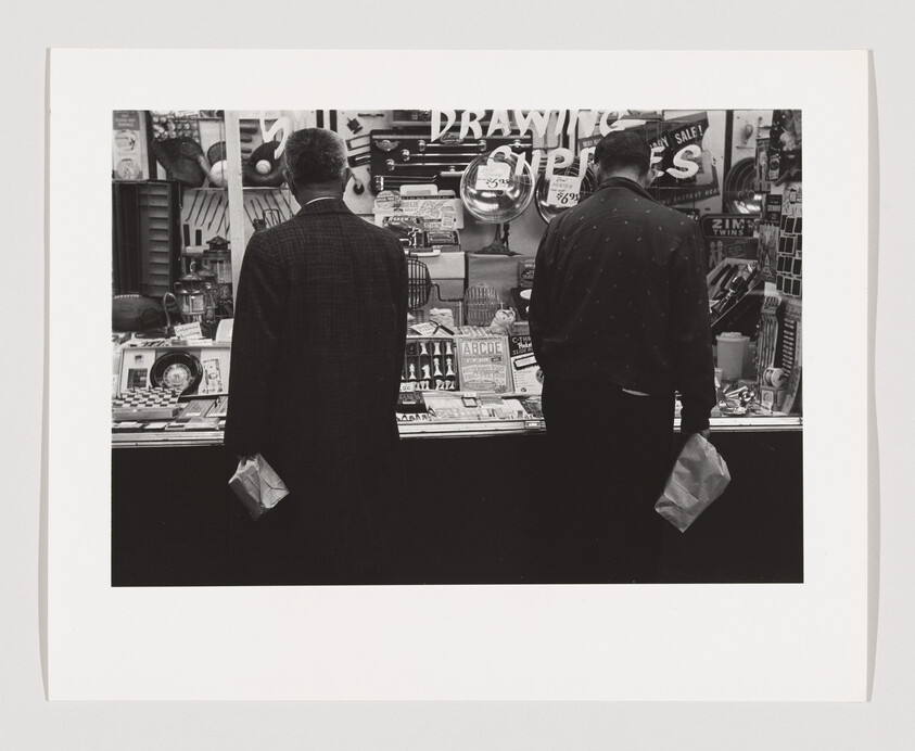 Two men stand with their backs to the camera, looking into a cluttered shop window, each holding a paper bag.