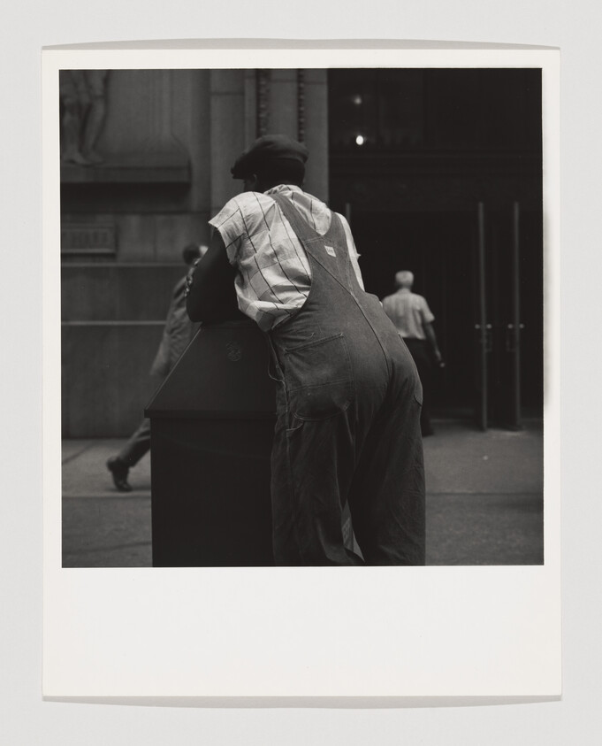 A black and white photograph capturing a person from behind as they lean over a dark box on the sidewalk. The person is wearing denim overalls, a plaid shirt, and a dark cap. In the background, other individuals are seen walking away, with the entrance of a building with classical architecture in the distance. The image has a white border framing it.