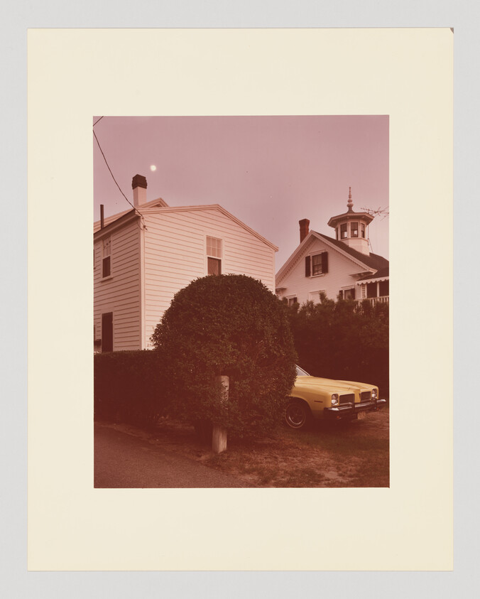 A yellow vintage car is parked beside a trimmed round hedge in front of two houses.