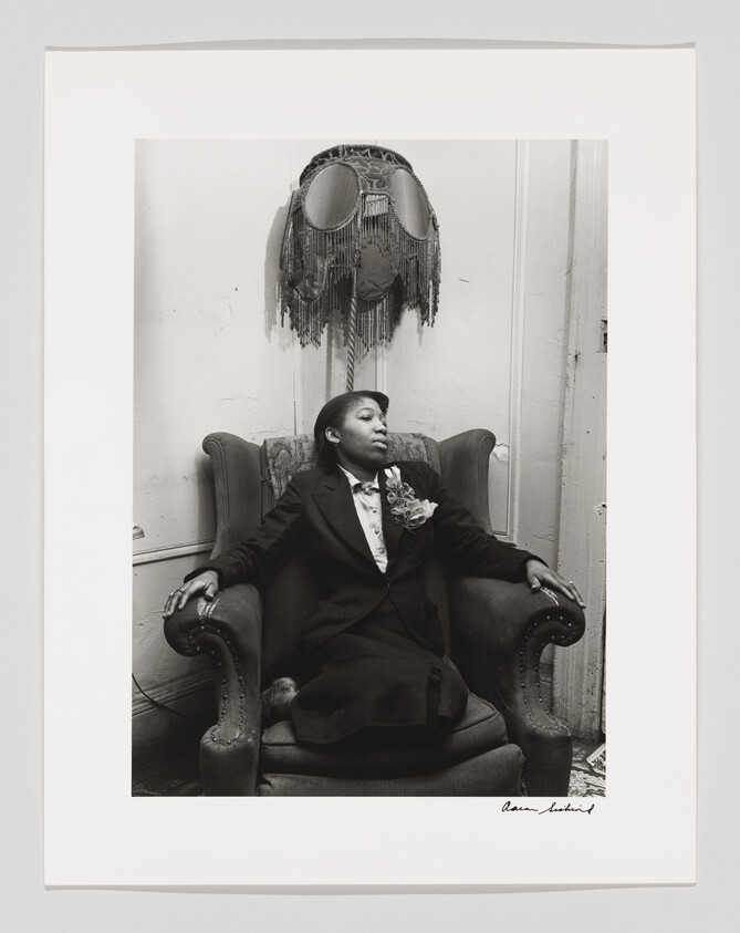 Young woman in a suit with a corsage reclines in an armchair, looking thoughtfully to the side.