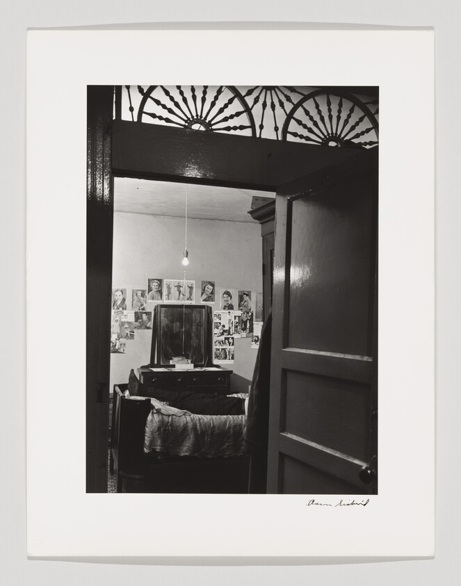 Open doorway reveals a small bedroom with a bed, dresser, mirror, and photos pinned to the wall.
