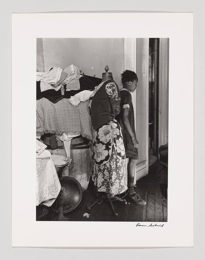 A black and white photograph depicting a young person standing in a cluttered room, looking out a window. They are wearing a patterned apron over a dress and socks with shoes. The room has a bed with disheveled linens, a wooden chair, and various household items scattered around, including a large metal bowl on the floor. The image has a signature on the bottom right.