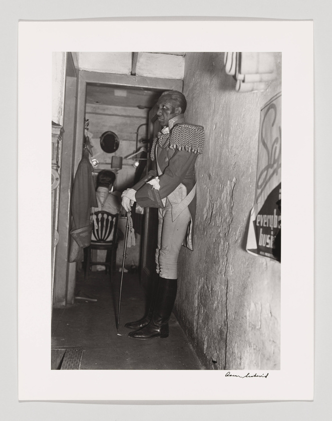 A black and white photograph depicting a person dressed in a historical military uniform with epaulettes, standing in a doorway. The individual is holding a cane and appears to be looking off to the side. In the background, there are chairs and a poster on the wall with the visible words "everybody's business." The photo has a signature at the bottom right corner.