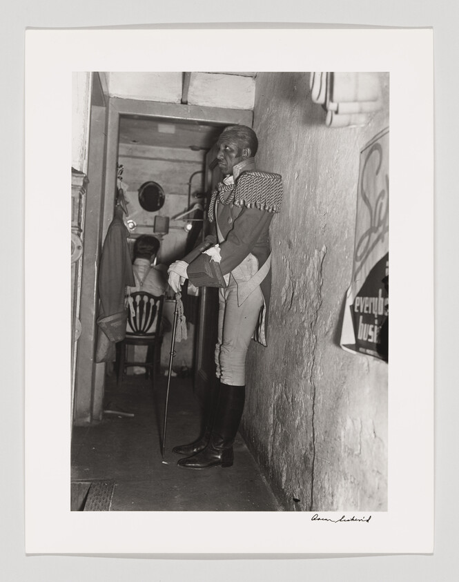 A man in a historical military uniform leans against a hallway wall holding a cane.