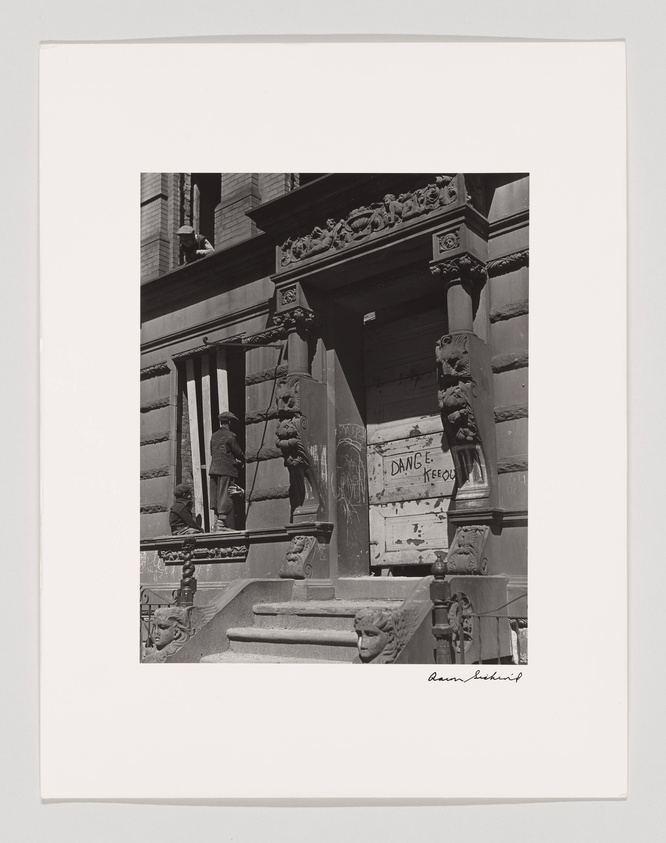 A black and white photograph capturing a moment on the steps of a building with ornate architectural details. Several children are seen playing on the staircase, which is adorned with classical sculptures and columns. A sign with the words "DANGER KEEP OUT" is posted on a boarded-up doorway, adding a sense of abandonment or disrepair to the scene. The image is signed by the photographer at the bottom right.