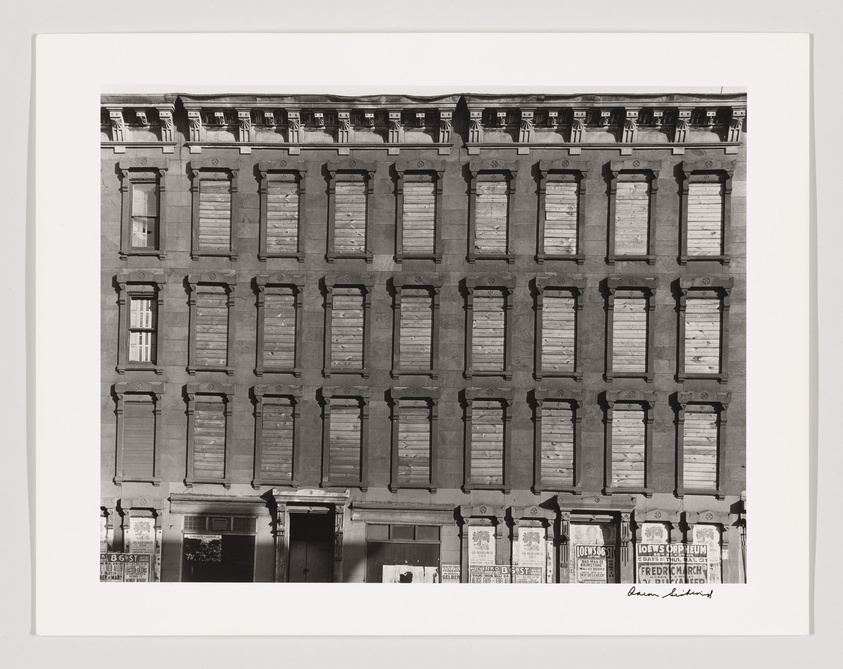 Black and white photograph of a symmetrical five-story building facade with boarded-up windows and a series of posters on the ground level. The building exhibits architectural uniformity with repetitive window patterns and a flat roofline. The photograph is framed with a white border and signed by the artist in the lower right corner.