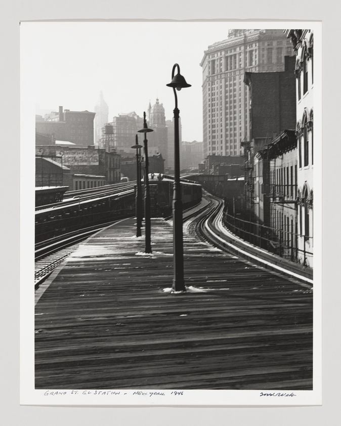 Black and white photograph of the Grand St. elevated train station in New York from 1946, showing curving tracks, a wooden platform, and vintage street lamps, with city buildings in the background shrouded in a light haze.