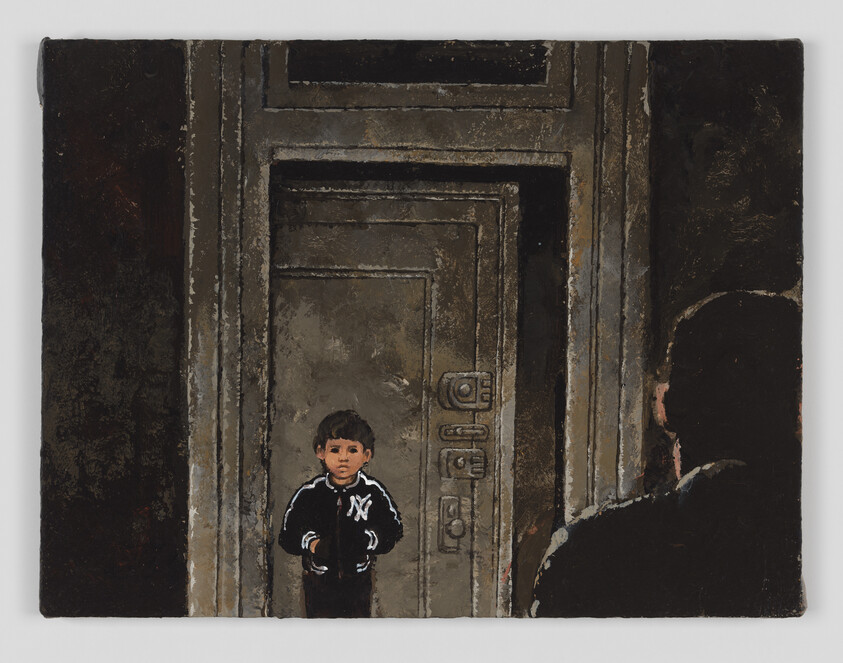 A young boy in a New York jacket stands before a large locked metal door while an adult watches.