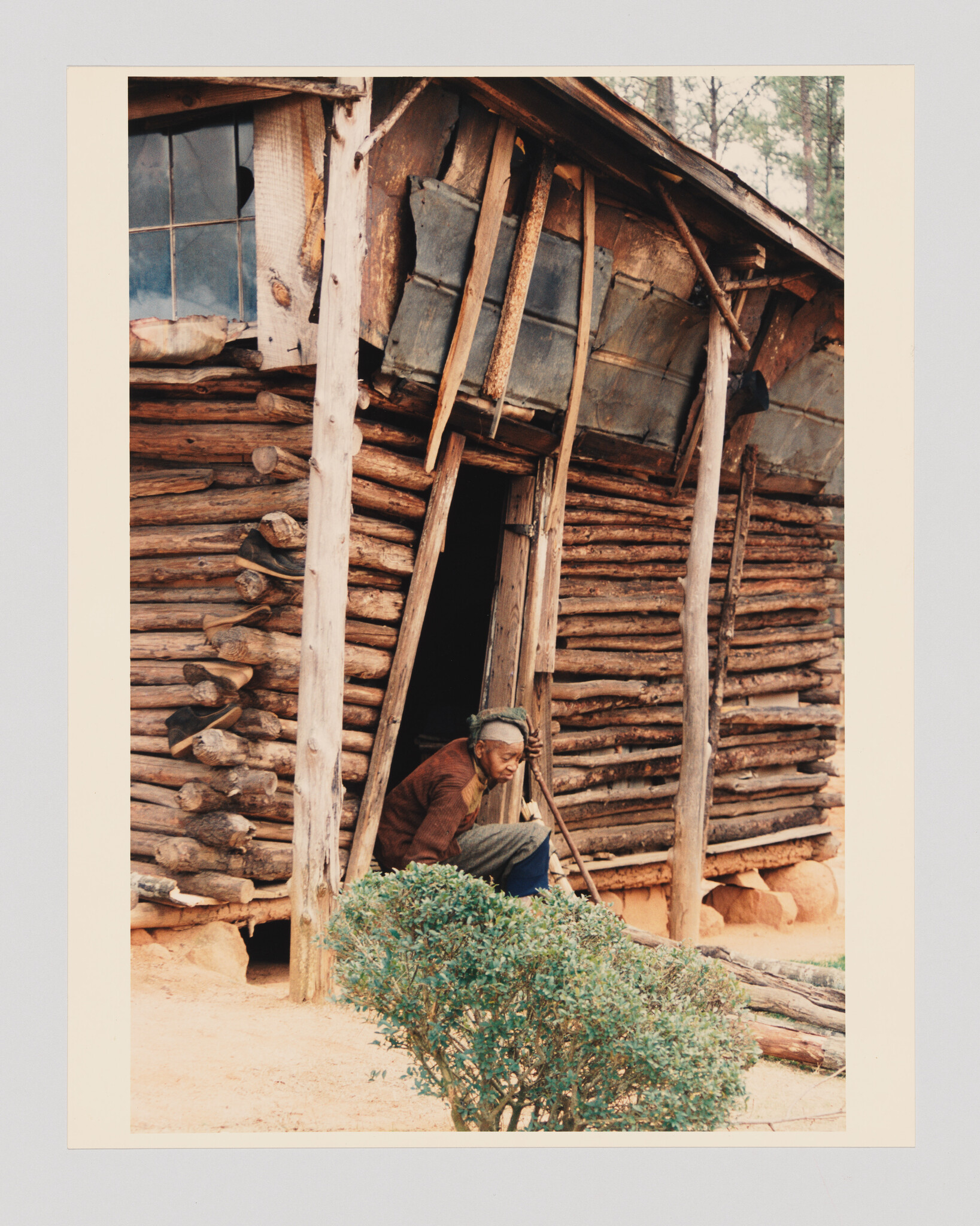 An elderly man sits in a leaning log cabin doorway holding a walking stick and looking down.