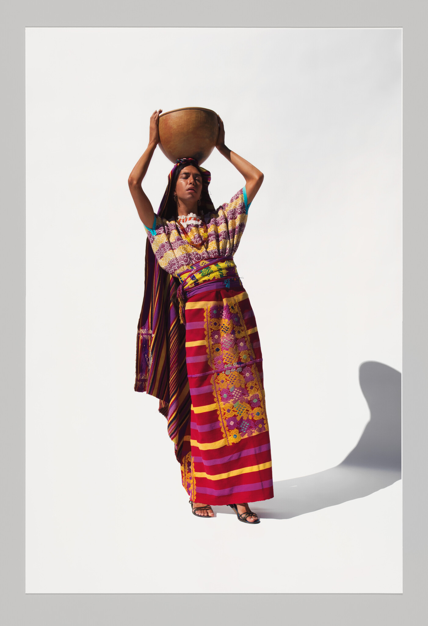 A woman in colorful traditional clothing balances a clay pot on her head with both hands.