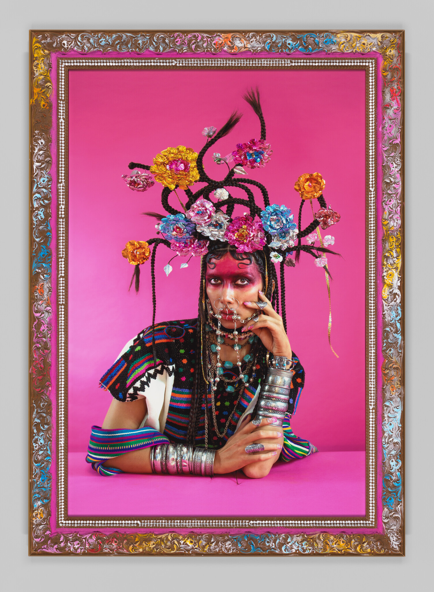 Person wearing braided floral headpiece and colorful clothing poses with chained face jewelry against pink.
