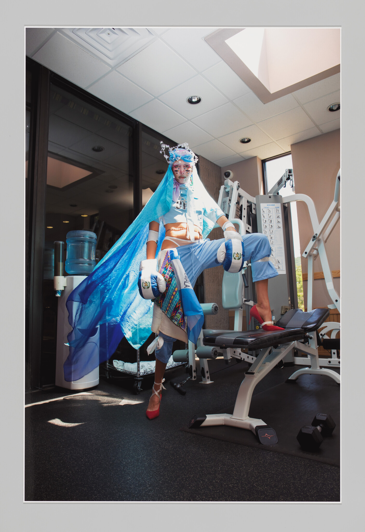 A person in an eclectic costume with a blue veil, face paint, and mismatched shoes is standing on one leg on a gym machine, striking a whimsical pose.