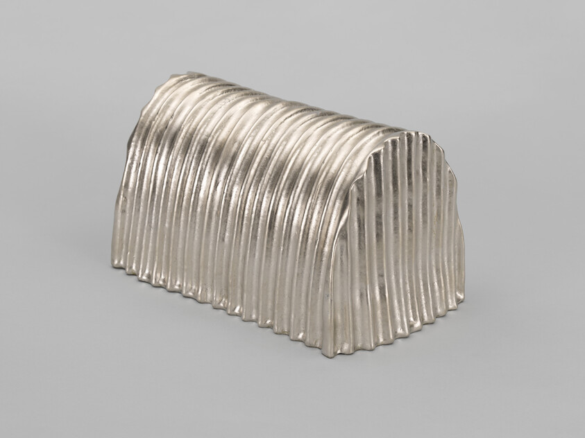 A silver corrugated object with a wavy texture and metallic sheen, resembling a miniature tunnel or a ridged architectural model, displayed against a solid gray background.