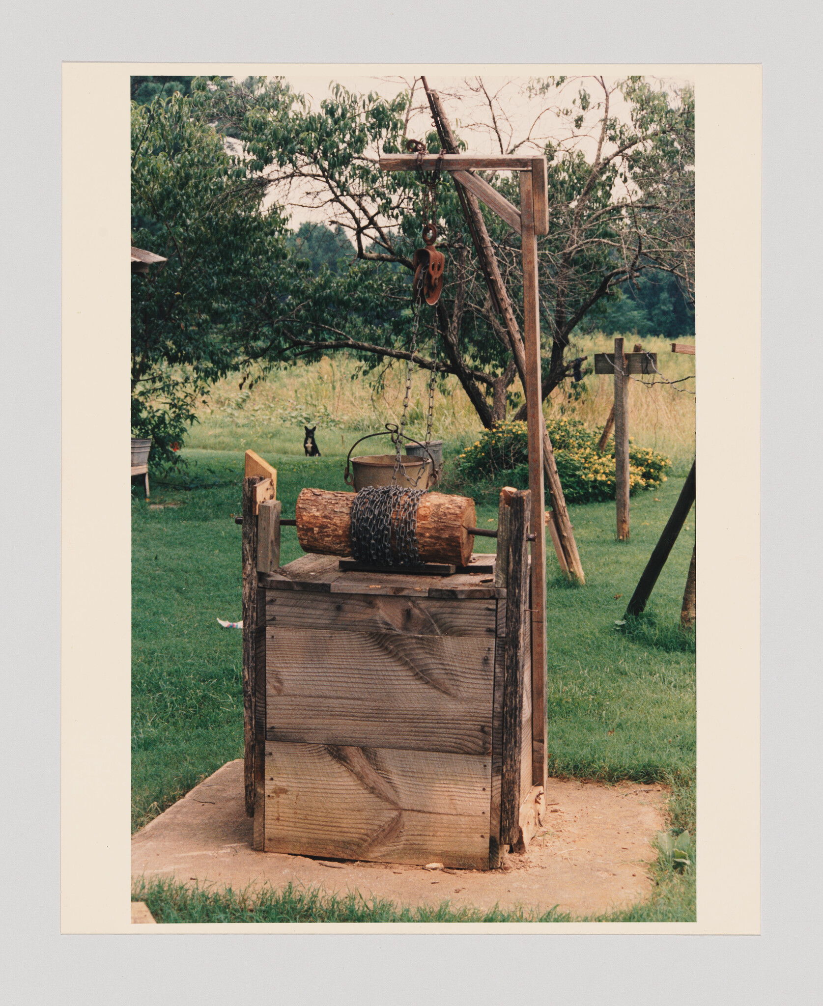 A rustic wooden well with a chain-wrapped log and hanging bucket sits in a grassy backyard.