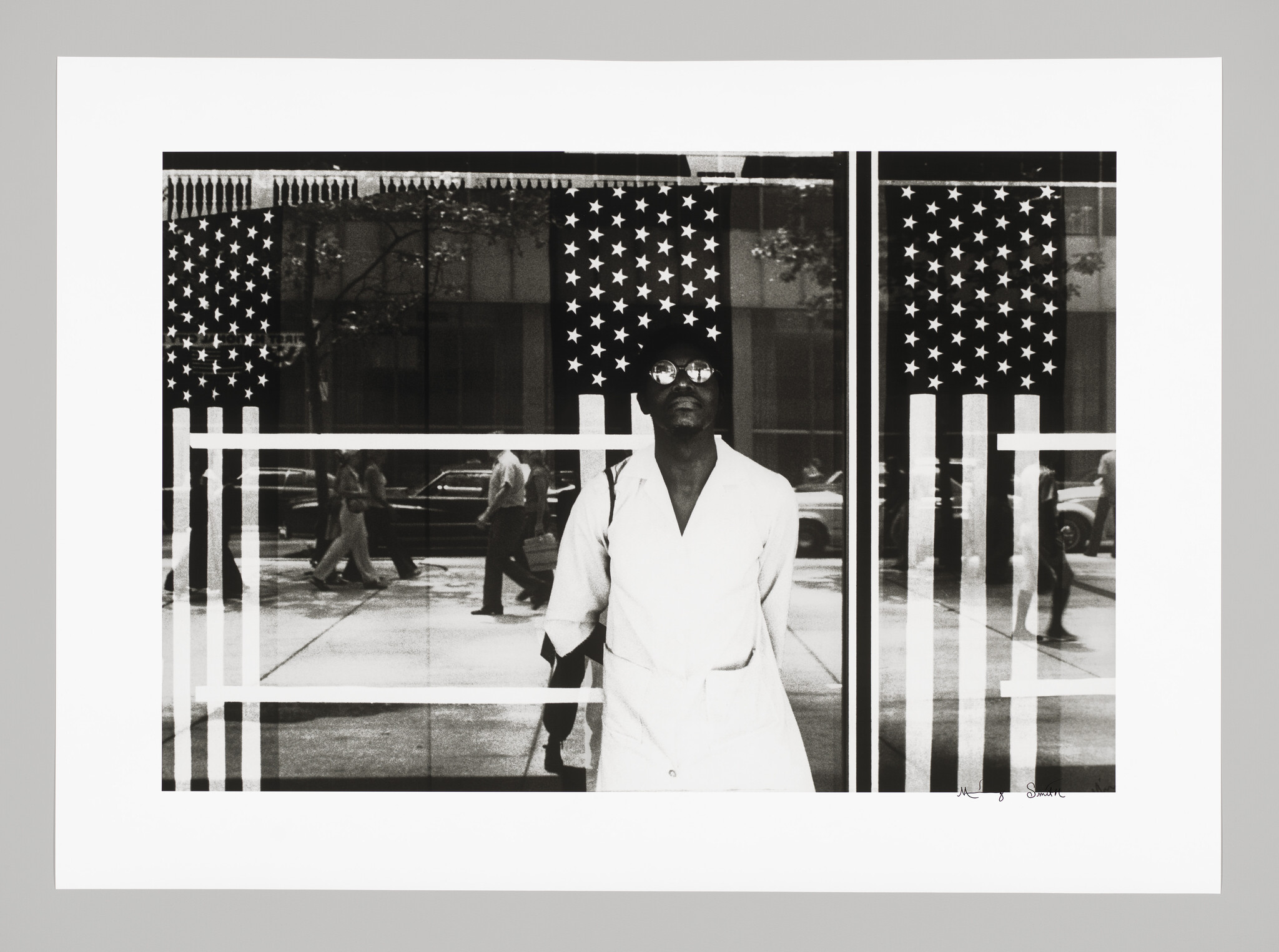 A man wearing sunglasses and a white coat stands before a glass window with American flag decals.