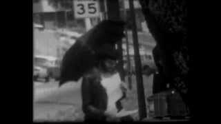 Black and white image of a person standing under an umbrella on a city street next to a street sign displaying the number 35.