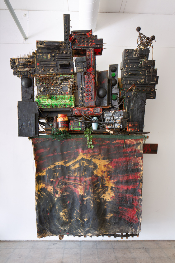 A complex assemblage sculpture made from various electronic components, circuit boards, and mechanical parts, mounted on a wall above a draped fabric with abstract red and black patterns. The sculpture has a dense, layered appearance with protruding elements and wires, and a few small green plants interspersed throughout.