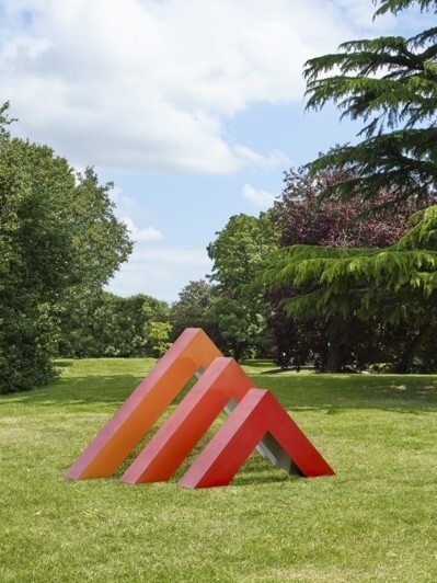 A vibrant orange and pink abstract sculpture consisting of slanted rectangular shapes is installed on a lush green lawn, with a backdrop of assorted trees under a partly cloudy sky.