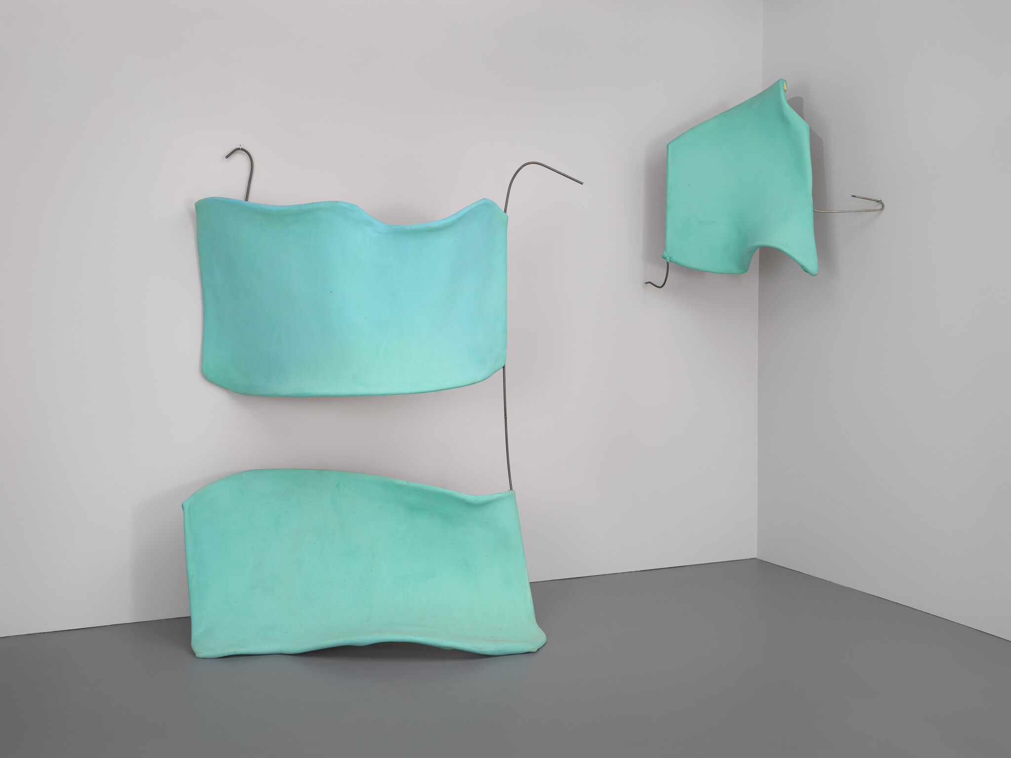 Three turquoise padded shapes hang from metal hooks on a bare gallery corner wall.