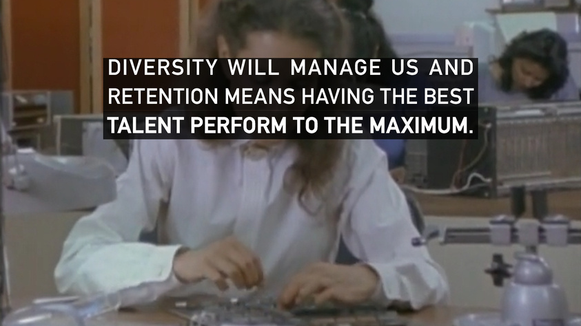 A still from a film or television show depicting an office setting with a person in the foreground typing on a keyboard and others working in the background. Overlaid text reads "DIVERSITY WILL MANAGE US AND RETENTION MEANS HAVING THE BEST TALENT PERFORM TO THE MAXIMUM."