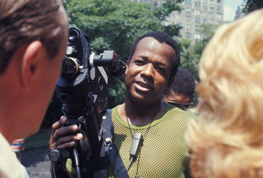 A man smiles as he looks into a film camera operated by another person whose back is to the camera. The man being filmed is wearing a green mesh top and has a microphone around his neck, suggesting an interview or filming in progress. The background is blurred with hints of greenery and buildings, indicating an outdoor setting.