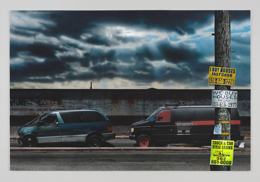 Two vans parked on a street with stormy skies above and various advertisements on a pole and graffiti on a wall in the background.