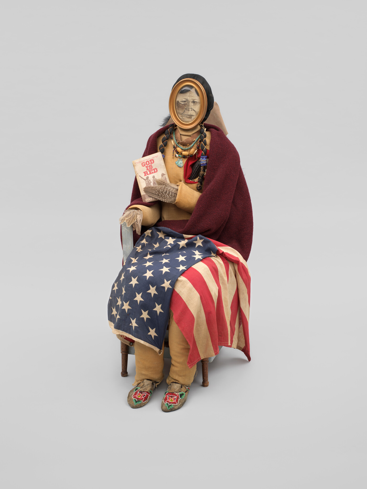 A seated Native American figure wrapped in an American flag and blanket holding the book "God Is Red."