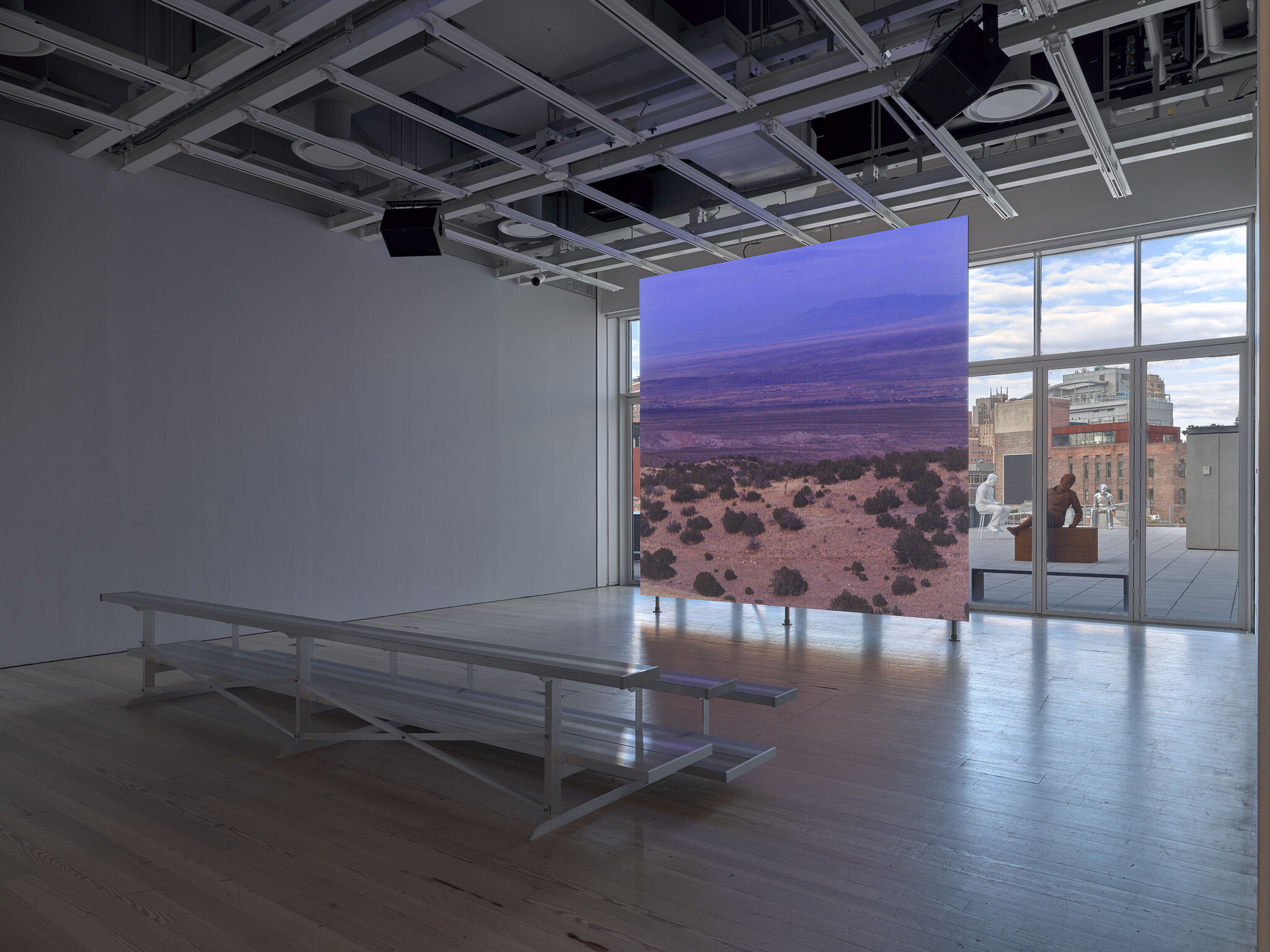 Modern gallery interior with a large landscape photograph on display, benches in the foreground, and cityscape visible through windows.