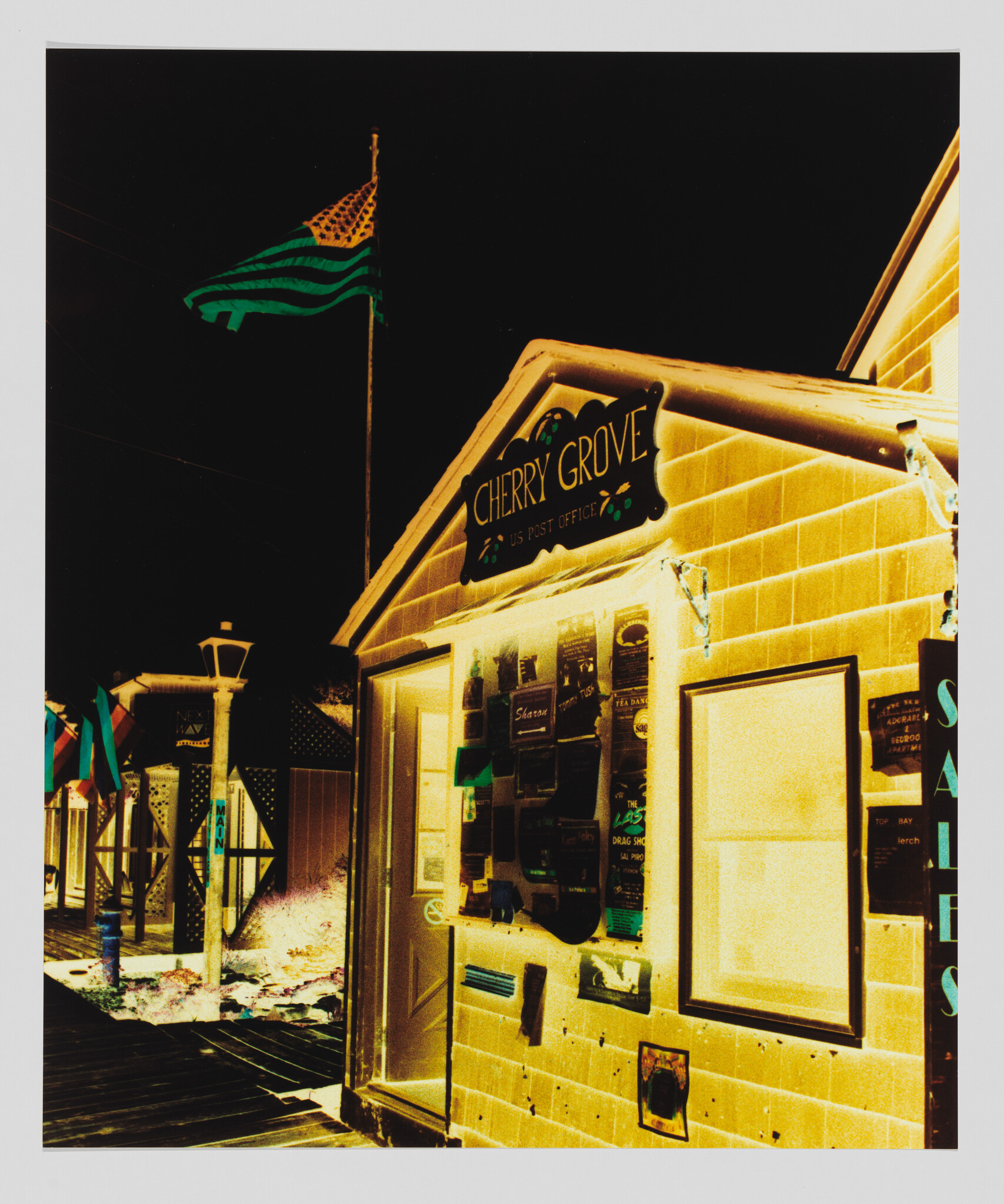 Small building with Cherry Grove sign and bulletin-covered wall under a flag at night.