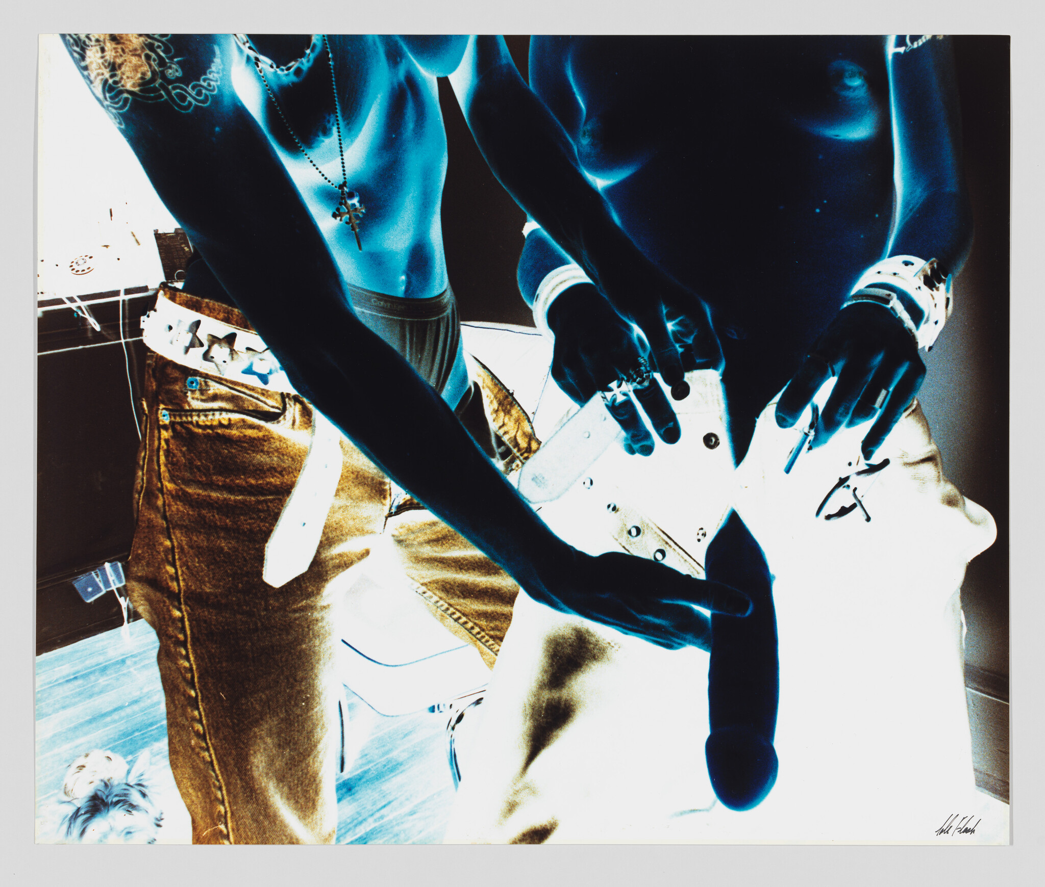 Two shirtless people wearing jeans adjust a large phallic sex toy attached to one person’s pants.