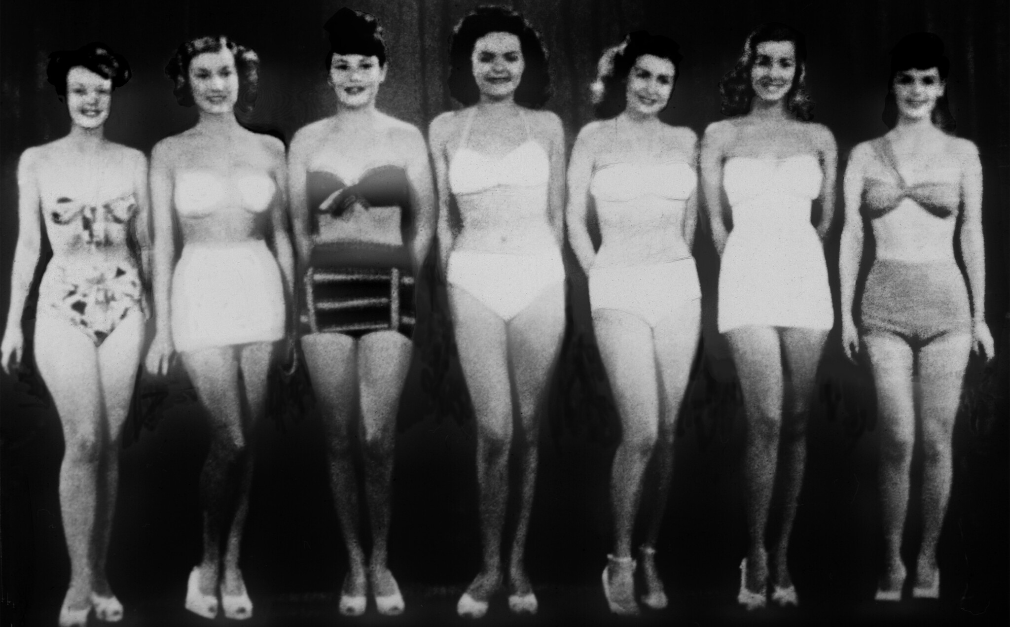 Seven young women stand in a line wearing vintage swimsuits and smiling for a contest.