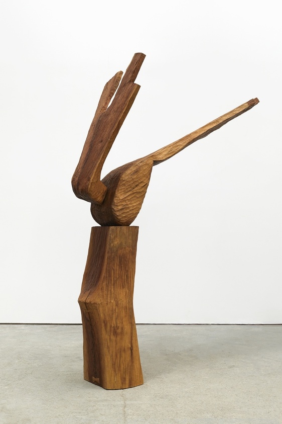 Wooden abstract sculpture resembling a bird with outstretched wings, standing on a textured base against a plain background.