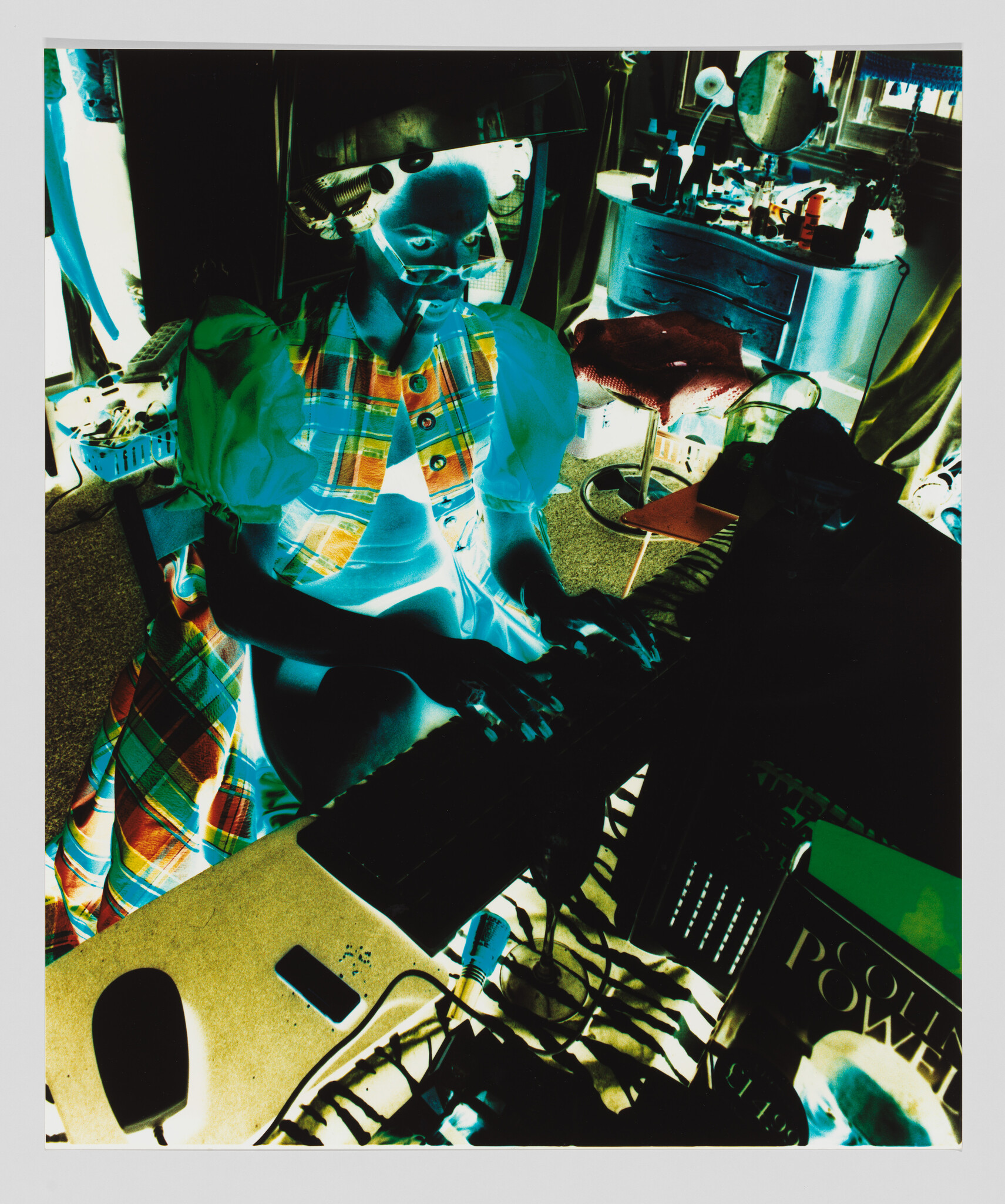 A person in a plaid dress sits at a keyboard, playing music in a cluttered room.