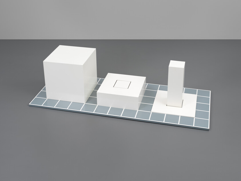 A minimalist composition featuring three white geometric shapes—a cube, a flat square with a smaller square indented, and a tall rectangular prism—arranged on a grid of light blue and white tiles, set against a grey background.