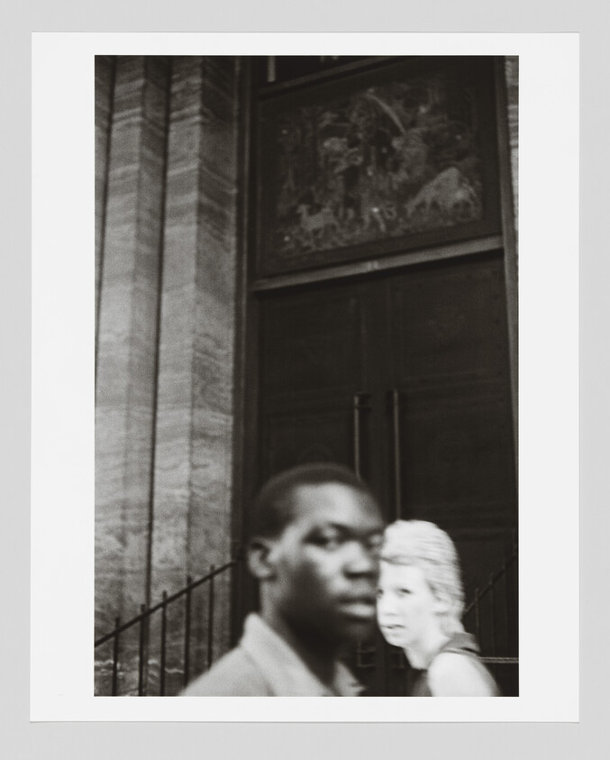 A blurred young man and a woman walk past a large dark doorway on a city street.