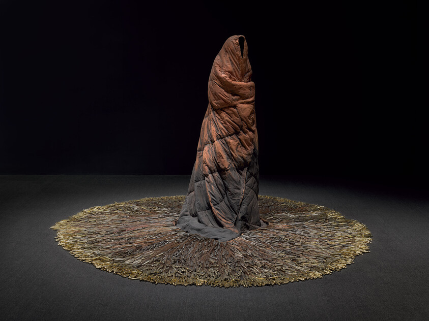 A tall rust-colored wrapped textile sculpture stands on a wide circular fringe of layered fabric.