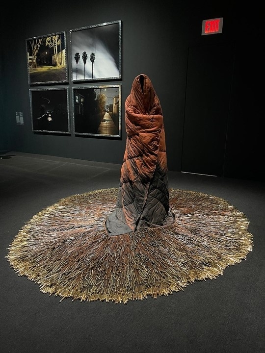 A sculpture resembling a cloaked figure stands on a circular base of keys, surrounded by framed photographs on a dark gallery wall.