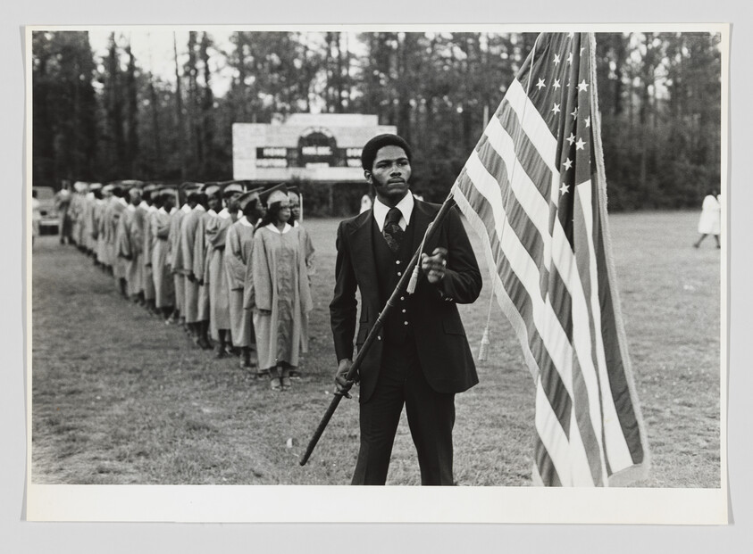 Young man in suit holds an American flag while graduates stand in line behind him.