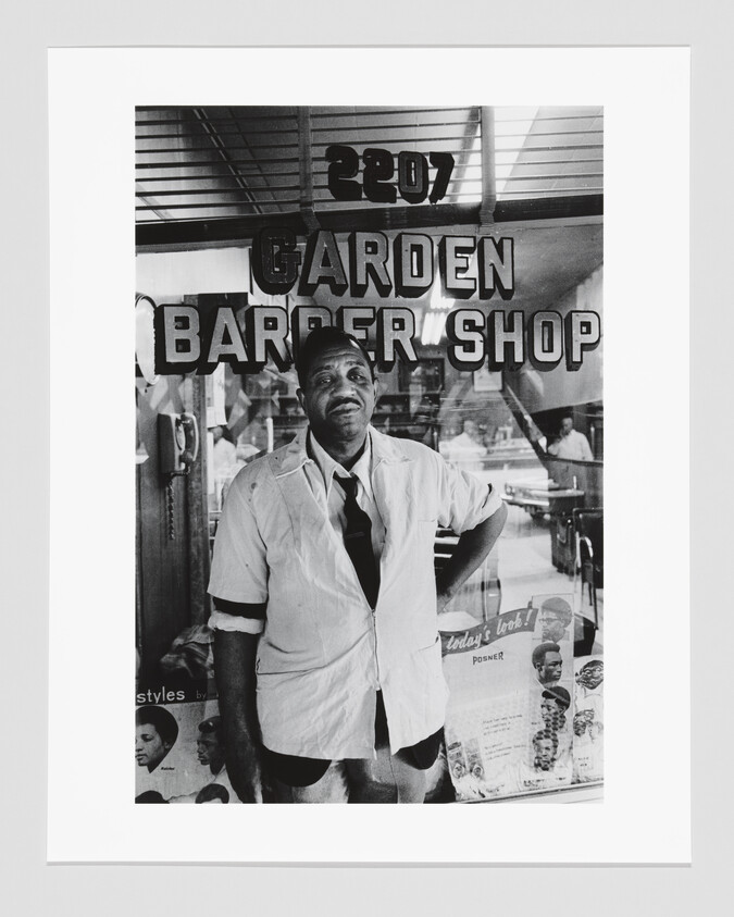 A barber stands with hand on hip in front of a window reading garden barber shop.