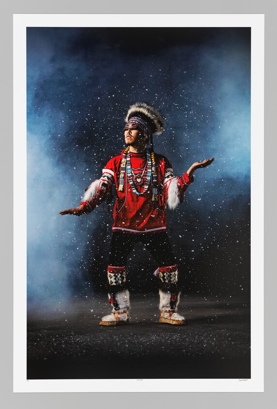 A person in traditional regalia stands with arms outstretched as snow falls around them.